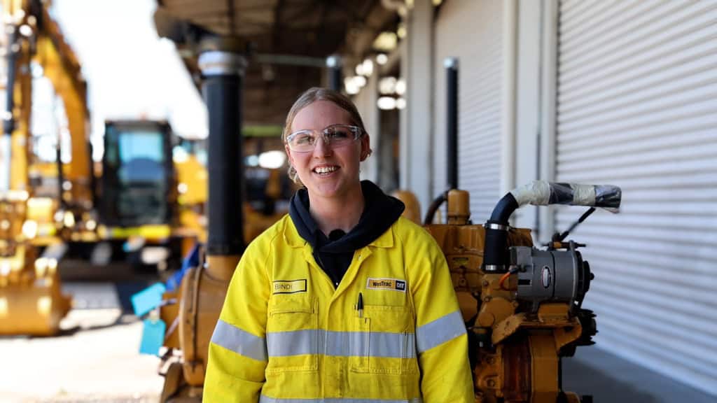 Bindi is a young female with blonde hair wearing her WesTrac PPE uniform while standing in front of a Cat engine.